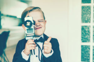Boy with blond hair shows a thumbs up sign while shooting a video with an old analog 8 mm retro camera. The boy wears a tuxedo with a bow tie.